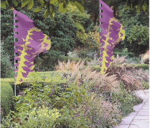Two flags, with shapes and printed color designs inspired by medieval Italian flags, flutering in a garden.